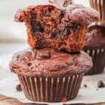 Two chocolate cottage cheese muffins are shown; one is whole and the other, with a bite taken out, rests on top, revealing a moist, chocolatey interior with visible chocolate chips. Both muffins sit on a wooden board.
