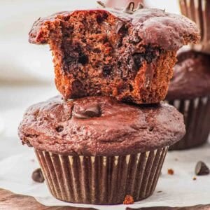 Two chocolate cottage cheese muffins are shown; one is whole and the other, with a bite taken out, rests on top, revealing a moist, chocolatey interior with visible chocolate chips. Both muffins sit on a wooden board.