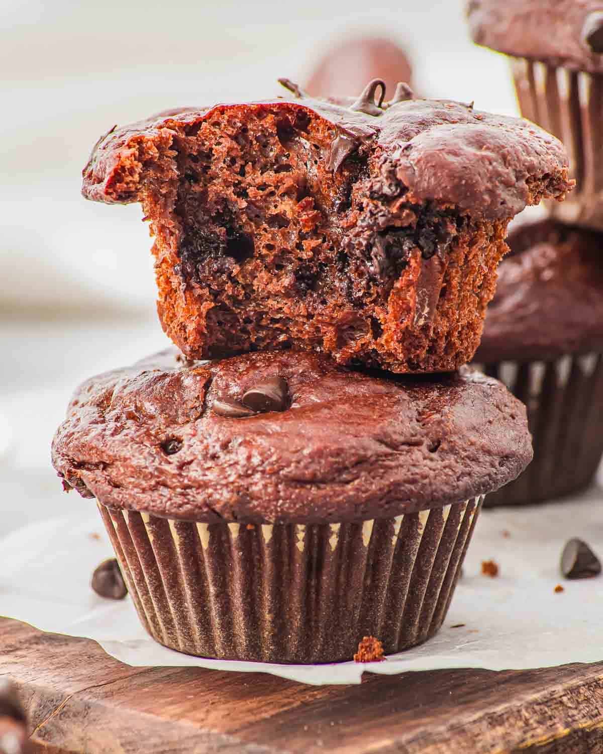 Two chocolate cottage cheese muffins are shown; one is whole and the other, with a bite taken out, rests on top, revealing a moist, chocolatey interior with visible chocolate chips. Both muffins sit on a wooden board.