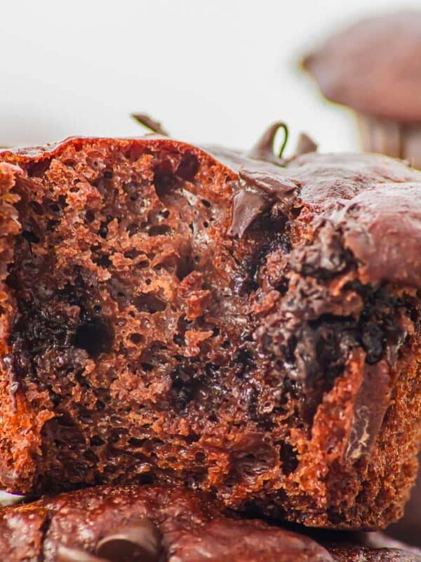 Close-up of a cottage cheese chocolate muffin with a bite taken out, showing a moist, gooey interior with melted chocolate chips. Another whole chocolate muffin is blurred in the background.