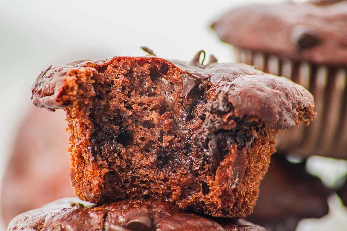 Close-up of a cottage cheese chocolate muffin with a bite taken out, showing a moist, gooey interior with melted chocolate chips. Another whole chocolate muffin is blurred in the background.