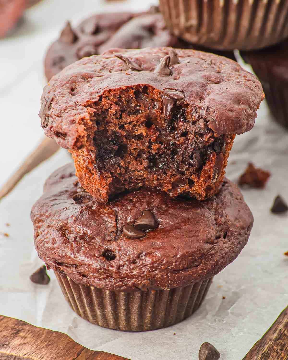 Two chocolate muffins are stacked, with the top muffin showing a bite taken out, revealing a moist, rich chocolate interior. Chocolate chips are scattered on and around the muffins.