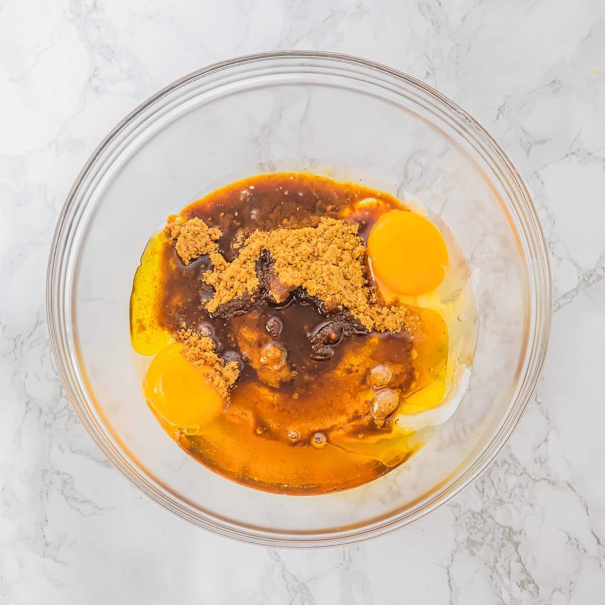 A glass bowl on a marble surface contains whipped cottage cheese, two eggs, coconut sugar, and vanilla extract, ready to be mixed for baking muffins.