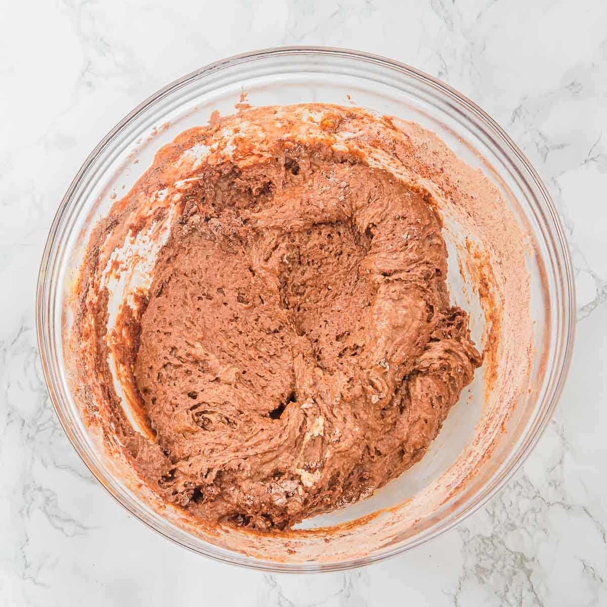A clear glass bowl containing thick chocolate muffin batter is placed on a white marble surface. The batter appears partially mixed with visible swirls and a slightly lumpy texture.