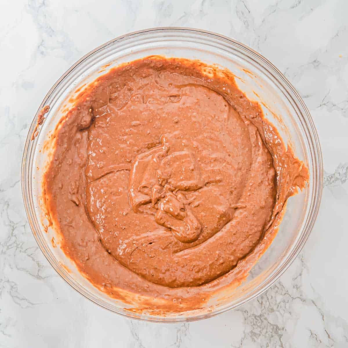 A glass mixing bowl filled with smooth chocolate muffin batter sits on a white marble surface.