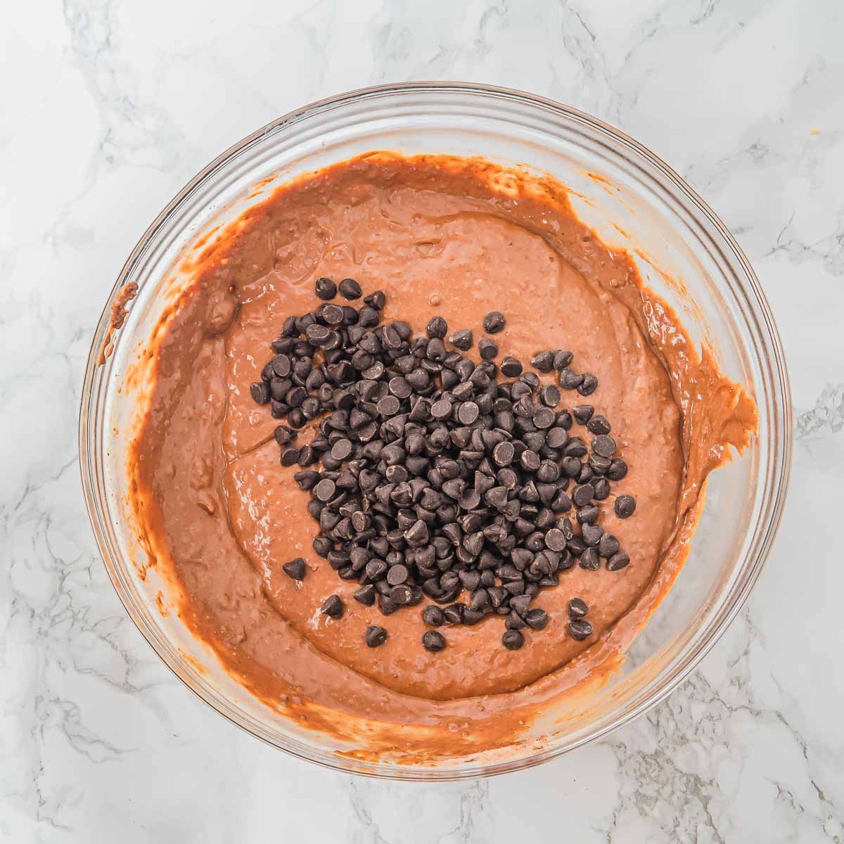 A glass bowl filled with chocolate batter sits on a marble surface, with a heap of chocolate chips added on top, ready to be mixed in.