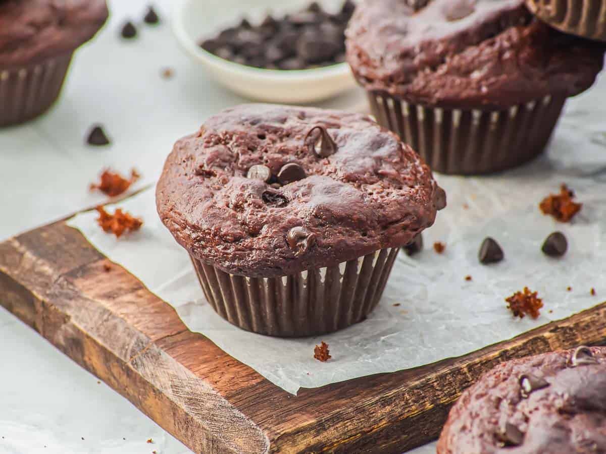 A close-up of a chocolate muffin made with cottage cheese and topped with chocolate chips is resting on parchment paper over a wooden board. More muffins, chocolate chips, and a bowl are visible in the background.