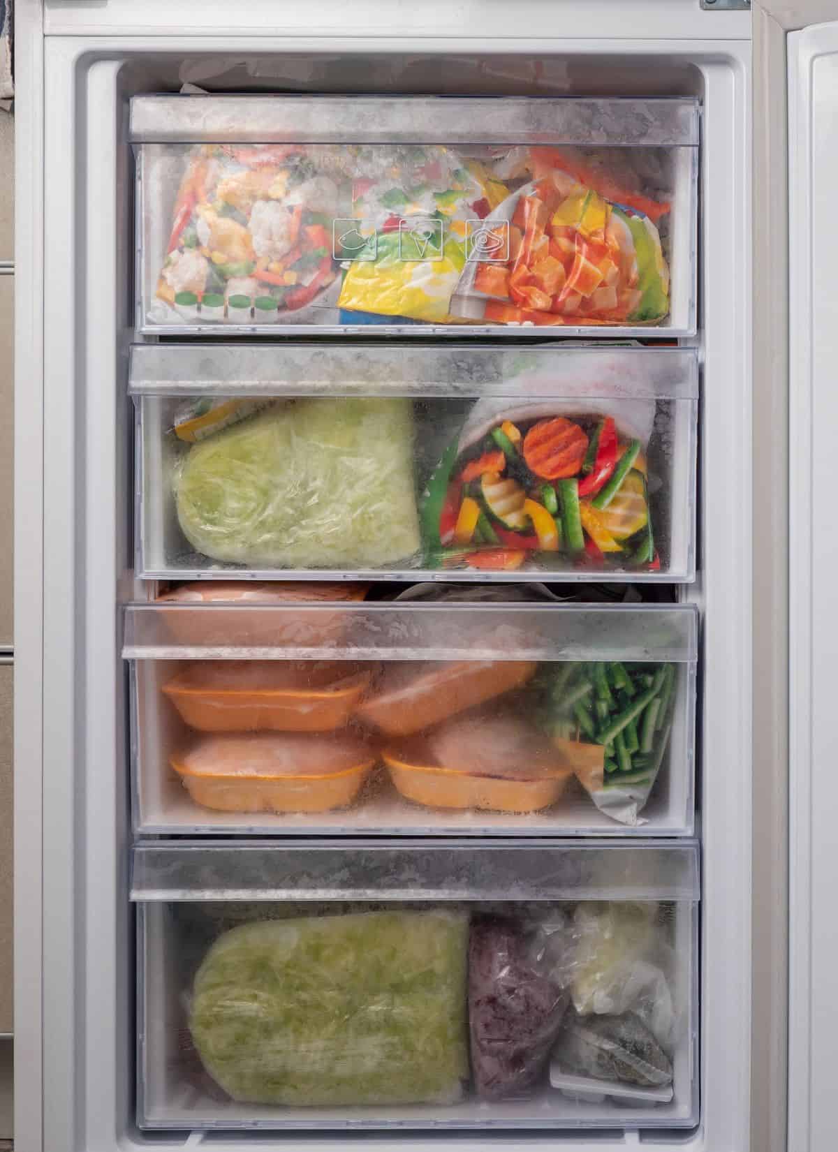 An open freezer with four drawers, each filled with various frozen foods, including vegetables, packaged meals, and plastic bags containing different items, all covered in a layer of frost.