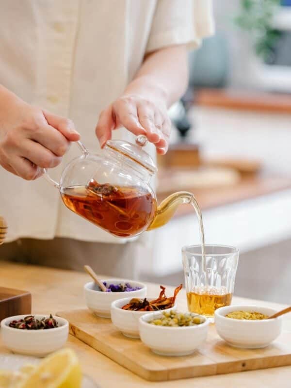 A person pours herbal tea from a glass teapot into a clear glass, showing how to make tea at home. Several small bowls with dried herbs and flowers are arranged on a wooden tray on the table.