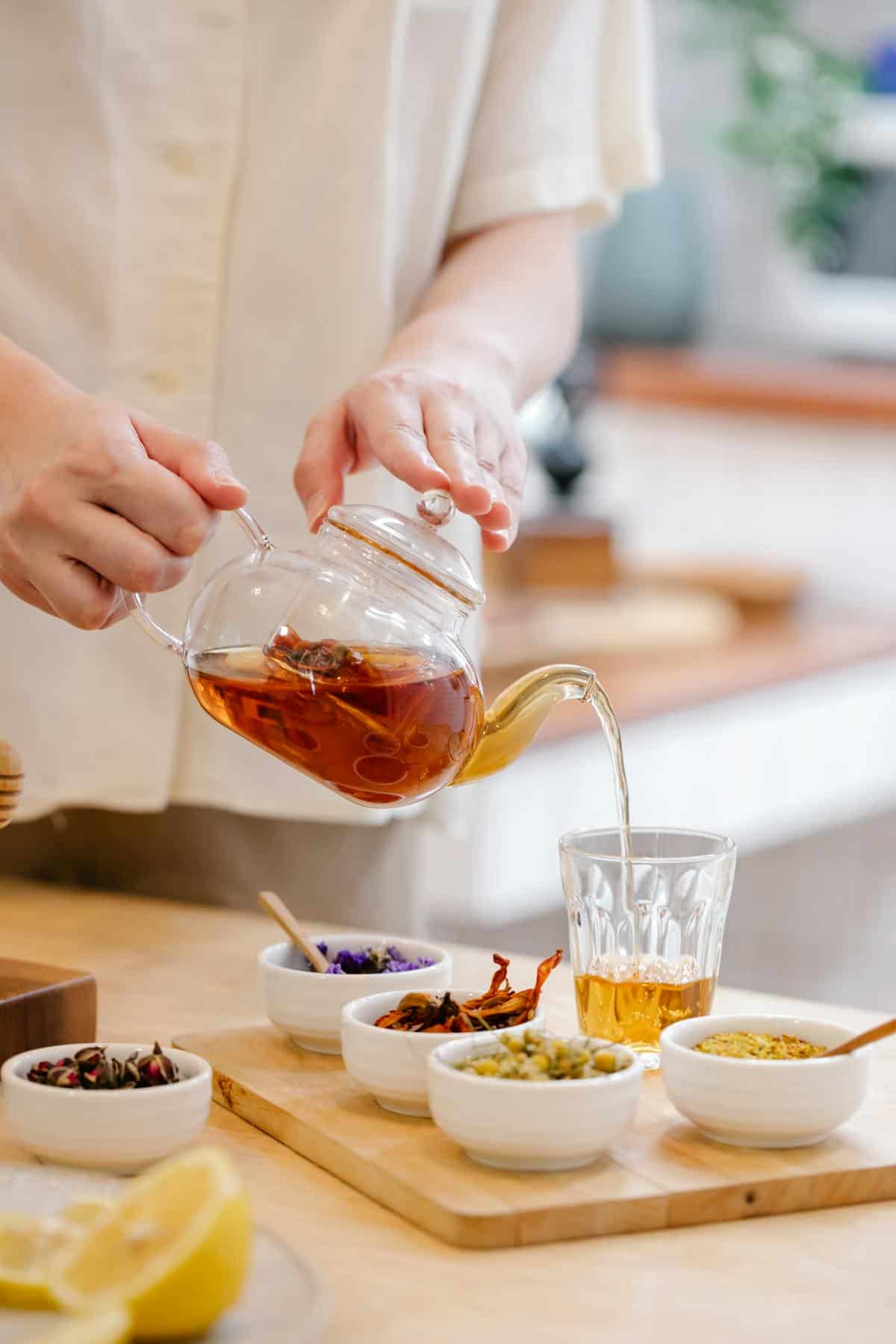 A person pours herbal tea from a glass teapot into a clear glass, showing how to make tea at home. Several small bowls with dried herbs and flowers are arranged on a wooden tray on the table.