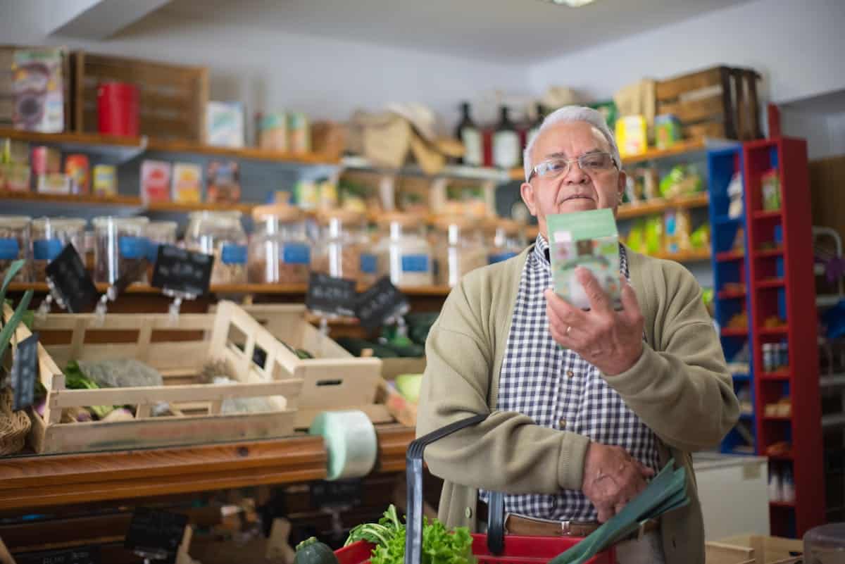 An elderly man with glasses stands in a grocery store, examining a product in his hand. He has a shopping basket with vegetables and is surrounded by shelves stocked with various food items.