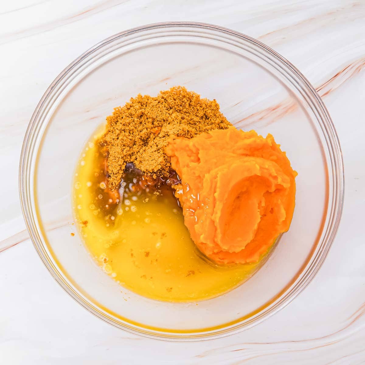 A glass bowl containing mashed sweet potato, melted butter, brown sugar, and vanilla extract on a light marble surface.
