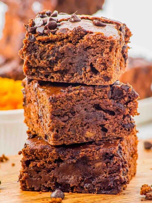 A close-up of three thick, fudgy chocolate brownies stacked on top of each other on a wooden surface, with visible chocolate chips and a gooey, moist texture.