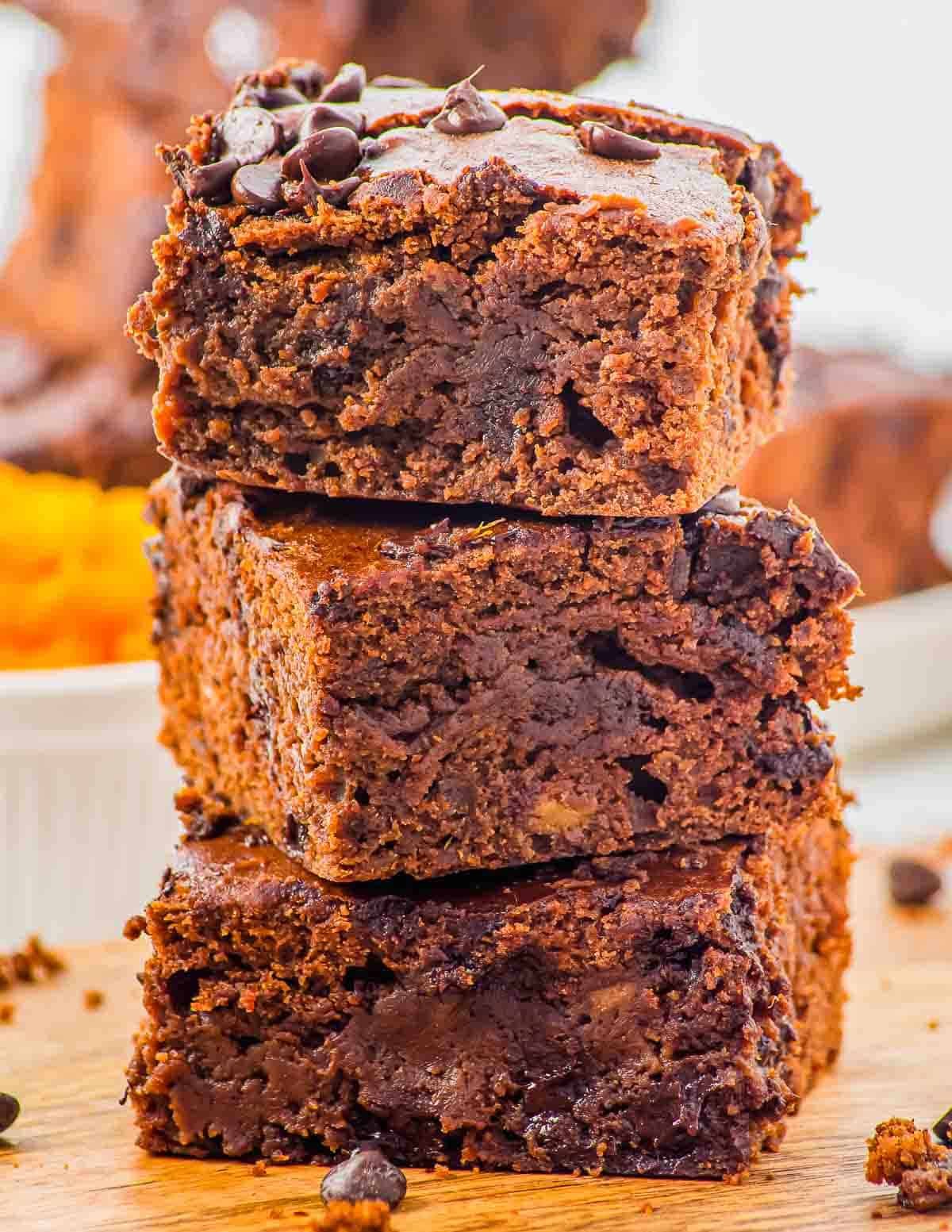 A close-up of three thick, fudgy chocolate brownies stacked on top of each other on a wooden surface, with visible chocolate chips and a gooey, moist texture.
