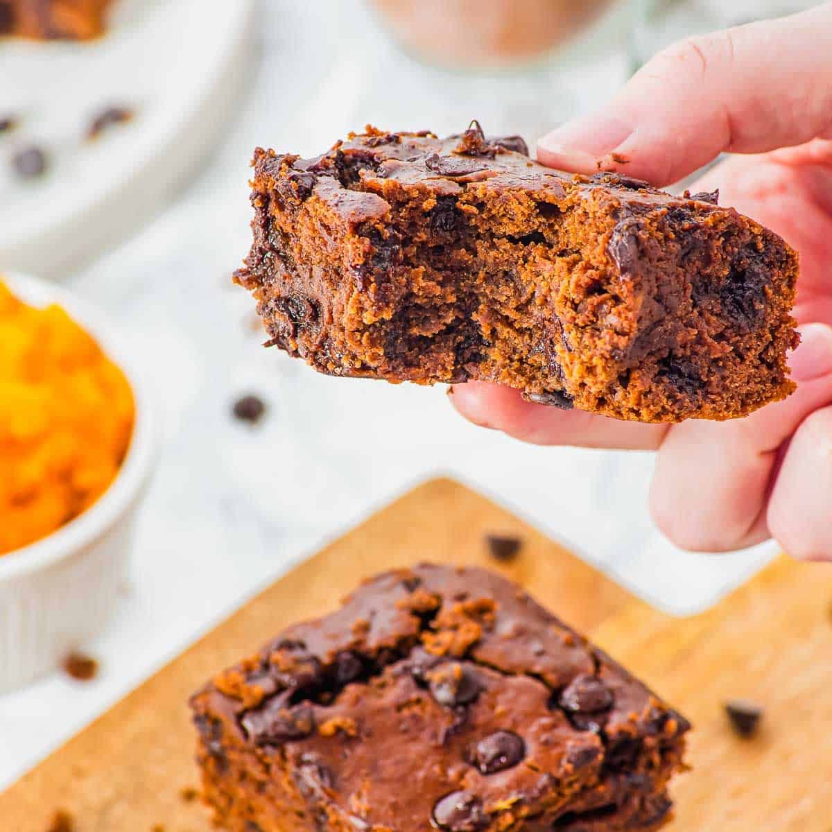 A hand holding a thick, moist chocolate chip sweet potato brownie with a bite taken out. More brownies rest on a wooden board below, with a small dish of pumpkin puree in the background.