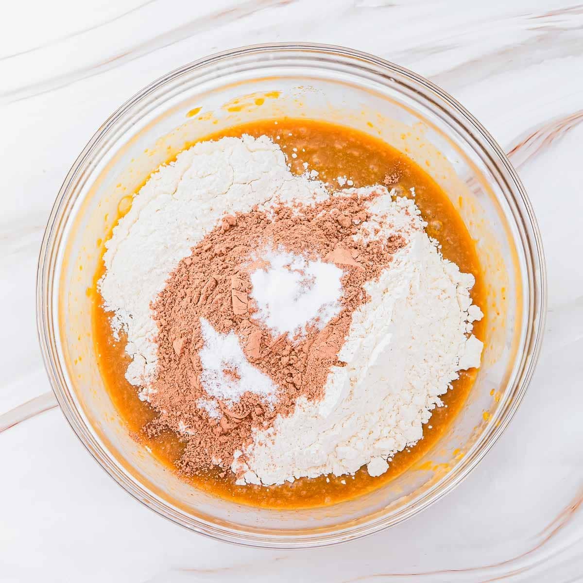 A glass bowl on a marble surface containing a mixture of wet ingredients with piles of flour, cocoa powder, and baking powder on top, ready to be mixed for baking.