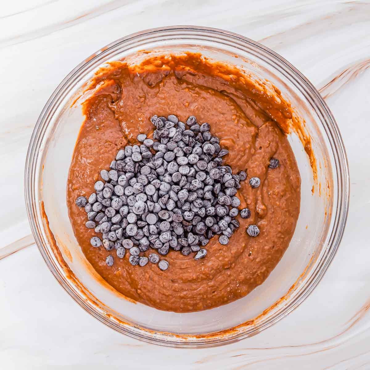 A glass bowl filled with sweet potato brownie batter topped with a pile of chocolate chips, sitting on a white marble surface.