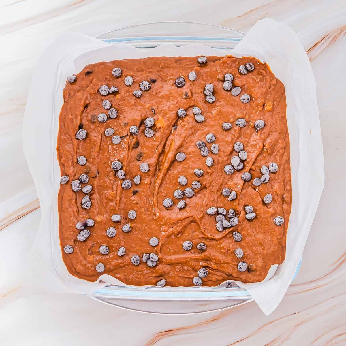 A square baking dish lined with parchment paper holds unbaked brownie batter topped with scattered chocolate chips, ready to be baked.