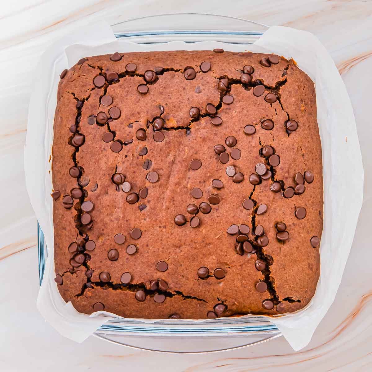 A square baking dish holding sweet potato brownies with a cracked top, baked in a parchment-lined glass dish, rests on a light-colored marble surface.