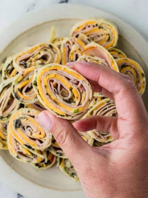 A hand holds a pinwheel-shaped appetizer made from a rolled tortilla with visible layers of ham, cheese, and herbs; several more pinwheels are arranged on a plate on a marble surface.