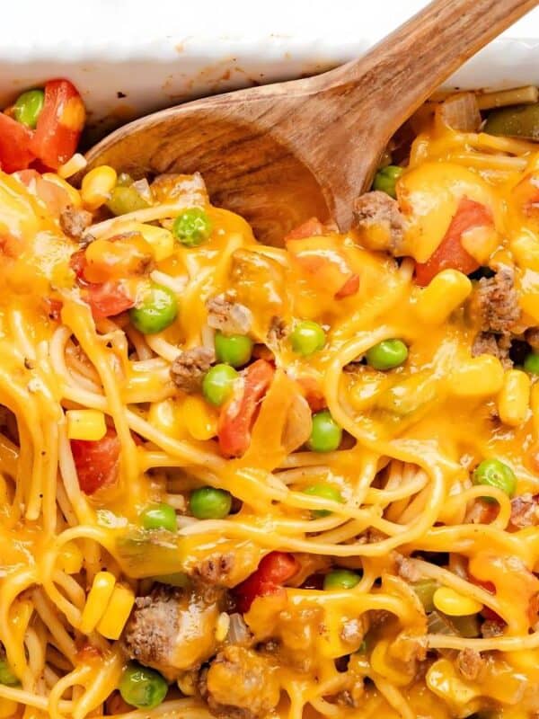 A casserole dish filled with baked spaghetti mixed with melted cheddar cheese, green peas, corn, diced tomatoes, and ground meat, with a wooden spoon scooping a portion.