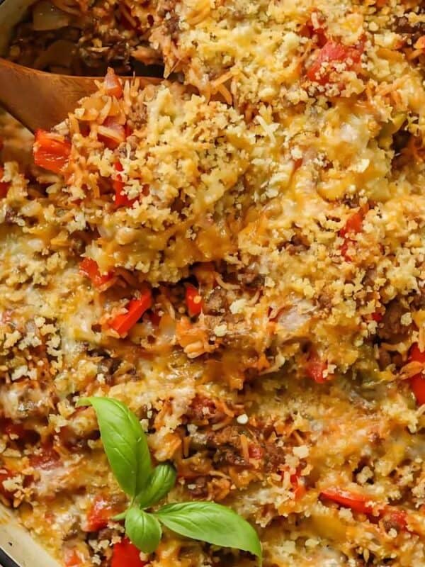 A close-up of a baked casserole with melted cheese, rice, red bell peppers, and a golden breadcrumb topping, garnished with fresh basil leaves in a large dish. A wooden spoon is scooping some of the casserole.