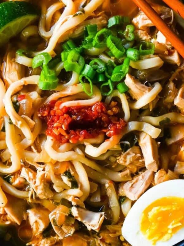 A bowl of noodle soup with shredded chicken, sliced green onions, chili paste, lime wedges, a soft-boiled egg, and a pair of chopsticks resting on the side.