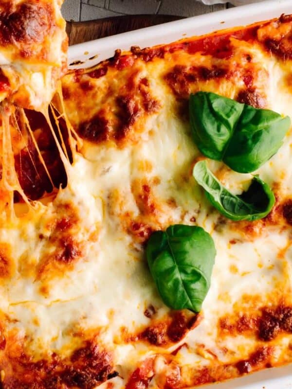 A baked lasagna in a white casserole dish, topped with melted cheese and fresh basil leaves. One square piece is being lifted, showing stretchy, melted cheese.