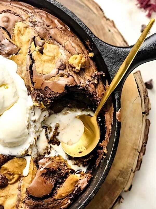 A cast iron skillet filled with swirled chocolate and peanut butter brownie, topped with scoops of vanilla ice cream, sits on a wooden board with a gold spoon and an empty white bowl nearby.