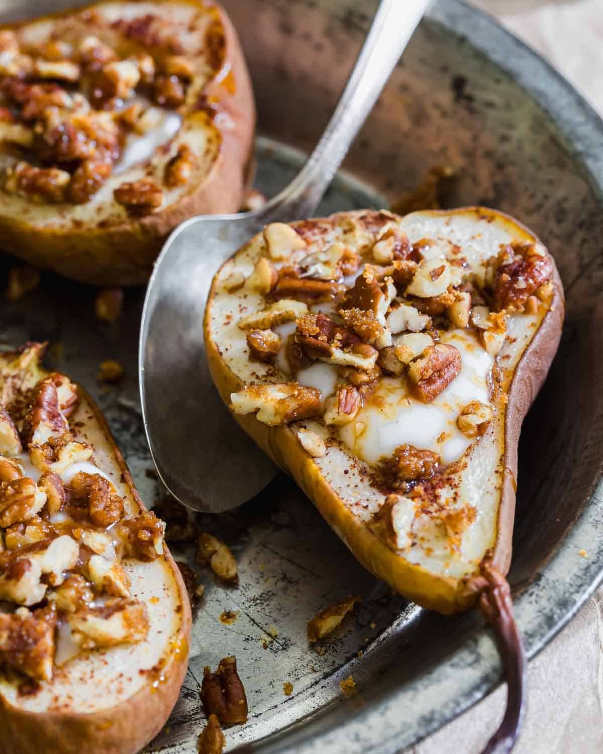 A baked pear half topped with chopped pecans and a drizzle of yogurt sits on a metal plate, next to a spoon. Another similarly topped pear half is partially visible in the background.