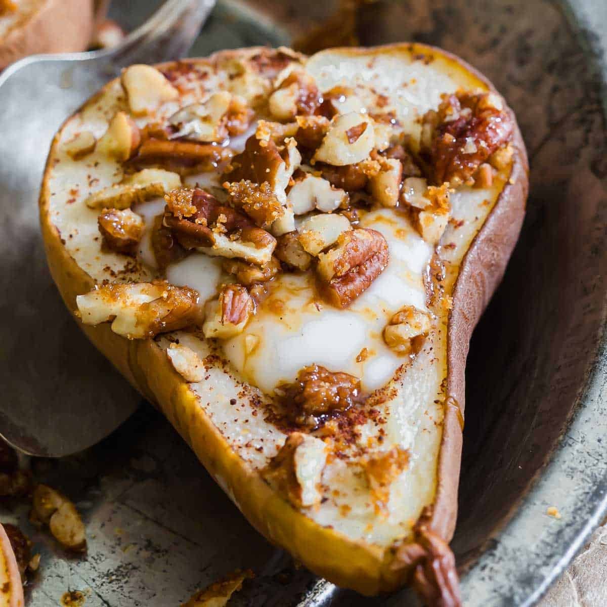 A baked pear half filled with creamy yogurt, topped with chopped nuts and a drizzle of syrup, served on a metal tray.