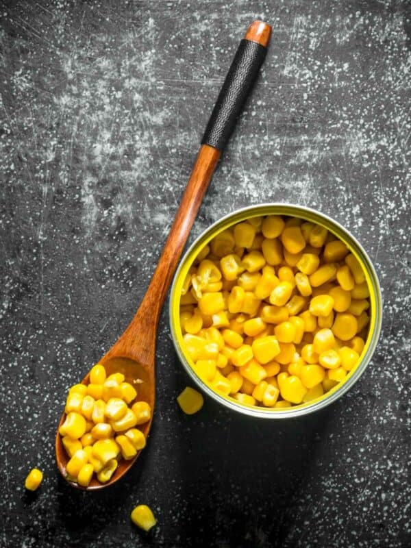 An opened can of yellow corn kernels sits on a dark textured surface next to a wooden spoon filled with corn, with a few kernels scattered nearby.