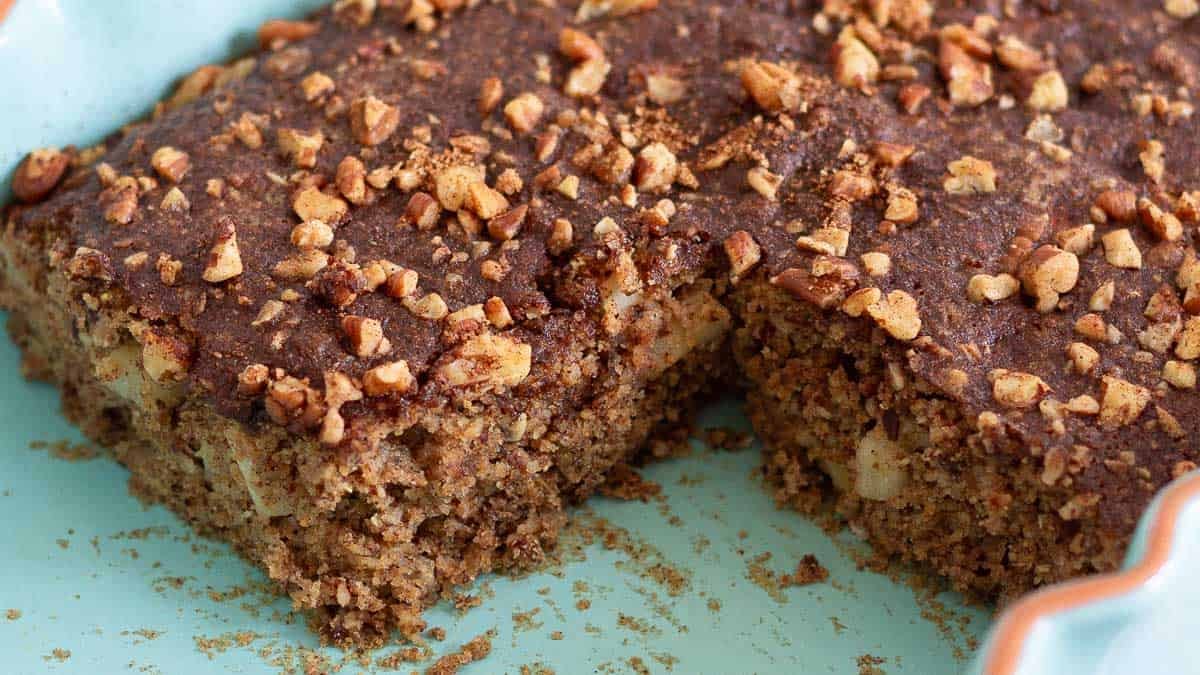 A close-up of a rectangular slice of cinnamon apple cake topped with chopped nuts, with one piece removed, revealing a moist and textured interior. The cake sits on a light blue plate.