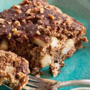 A close-up of a slice of apple cinnamon cake with chopped nuts on top, resting on a blue-green plate. A fork is positioned next to the cake, partially cutting into it.