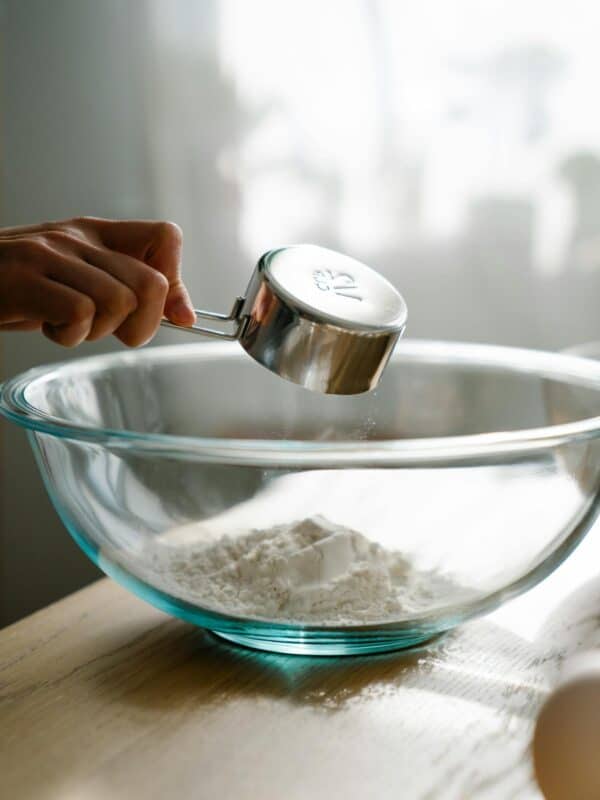 A hand pours flour from a metal measuring cup into a large clear glass mixing bowl on a wooden table, with soft daylight illuminating the scene.