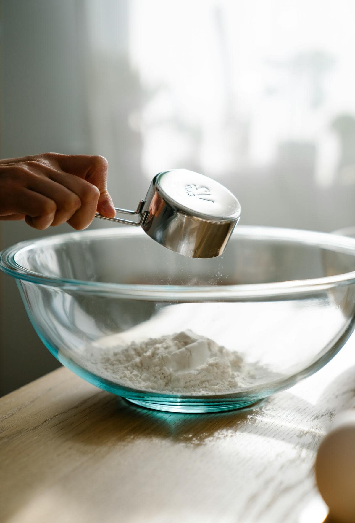 A hand pours flour from a metal measuring cup into a large clear glass mixing bowl on a wooden table, with soft daylight illuminating the scene.