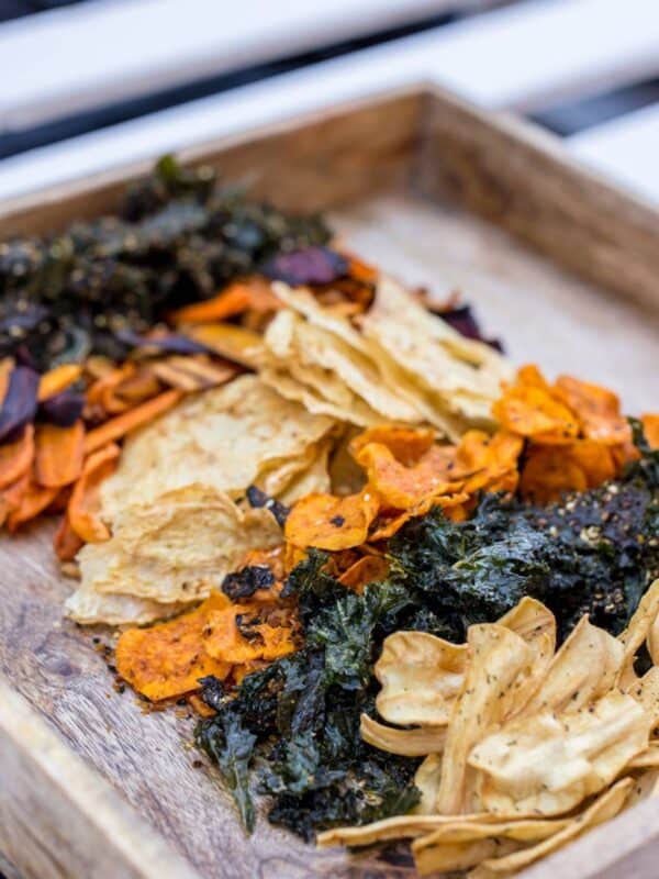 A wooden tray filled with an assortment of dehydrated vegetable chips, including kale, carrots, and possibly sweet potato, arranged in colorful, uneven piles.