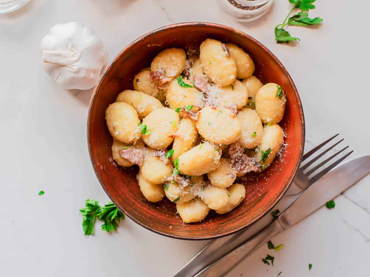 A bowl of gnocchi topped with grated cheese and herbs sits on a white table, surrounded by garlic, chopped parsley, a fork, and a knife.