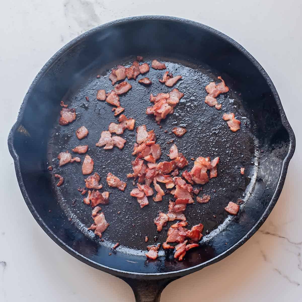 Chopped pieces of cooked bacon are scattered in a black cast iron skillet on a white countertop.