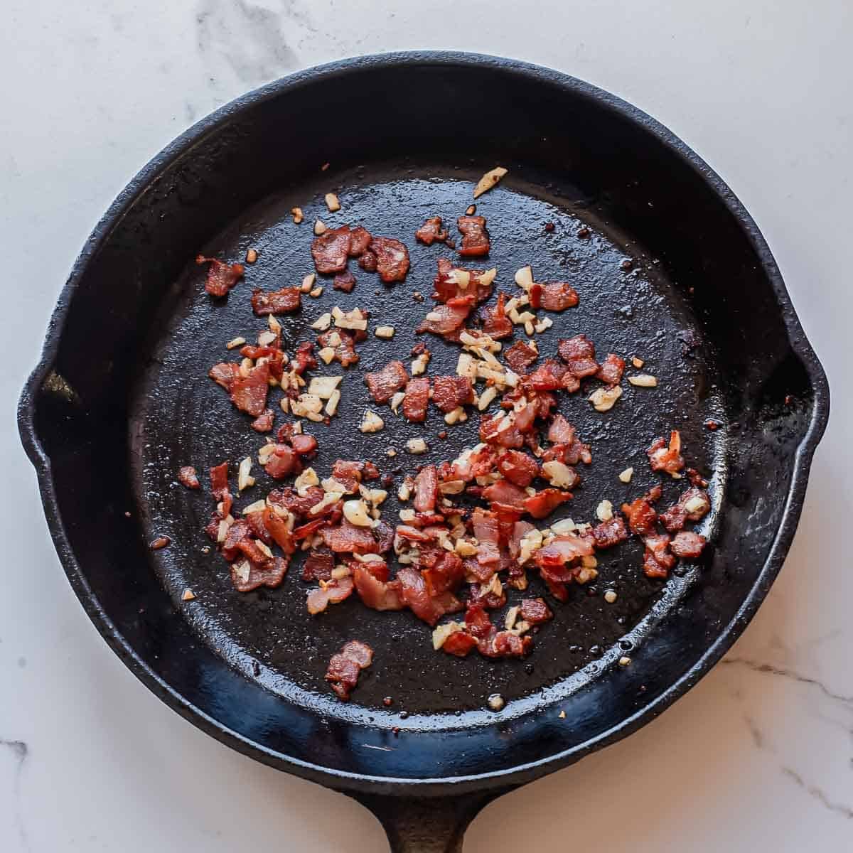 A black cast iron skillet containing small pieces of browned bacon and chopped garlic sits on a light-colored countertop.