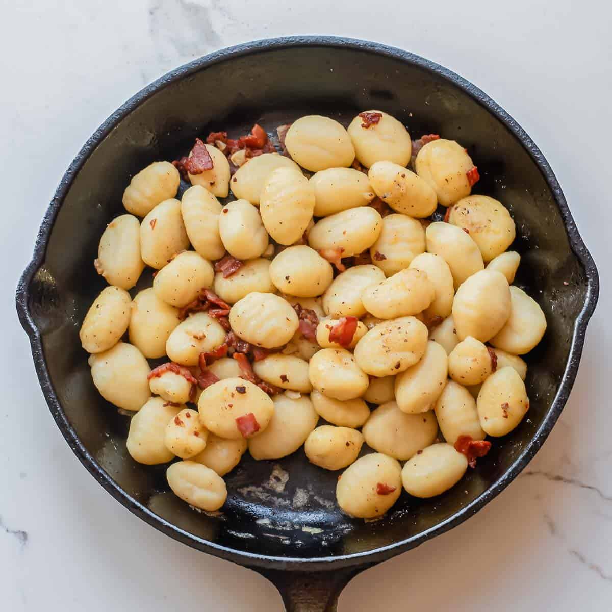 A cast iron skillet filled with cooked gnocchi and small pieces of bacon, sits on a white marble surface.