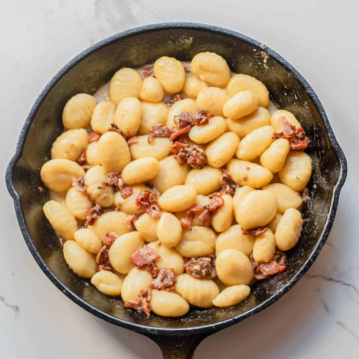 A black skillet filled with cooked gnocchi and pieces of crispy bacon in a creamy carbonara sauce sits on a white marble surface.