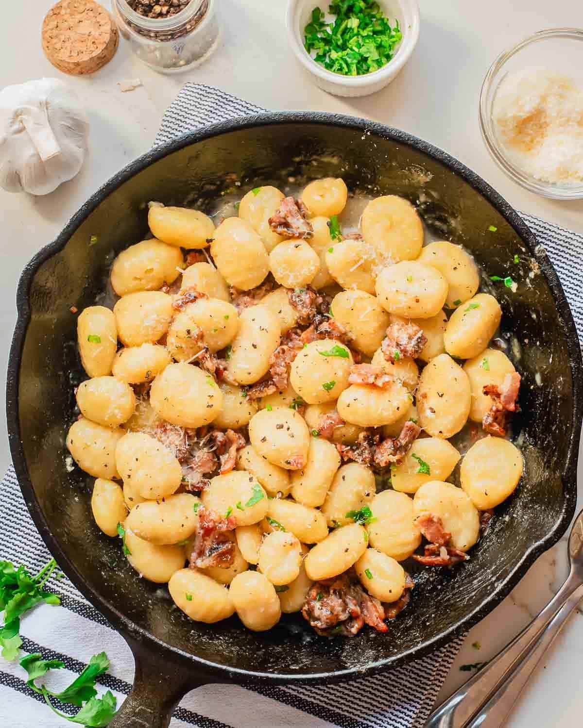 A skillet filled with gnocchi carbonara with pieces of bacon, garnished with chopped parsley. Surrounding the skillet are bowls of grated cheese, chopped herbs, and a bulb of garlic on a striped kitchen towel.