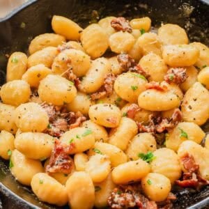 A skillet filled with creamy gnocchi mixed with pieces of crispy bacon, garnished with chopped parsley and grated cheese.