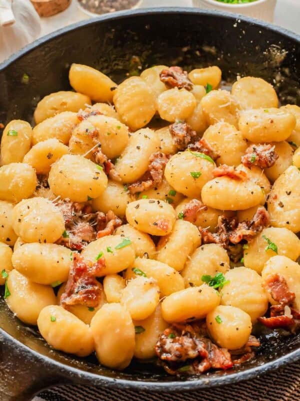 A skillet filled with creamy gnocchi mixed with pieces of crispy bacon, garnished with chopped parsley and grated cheese.