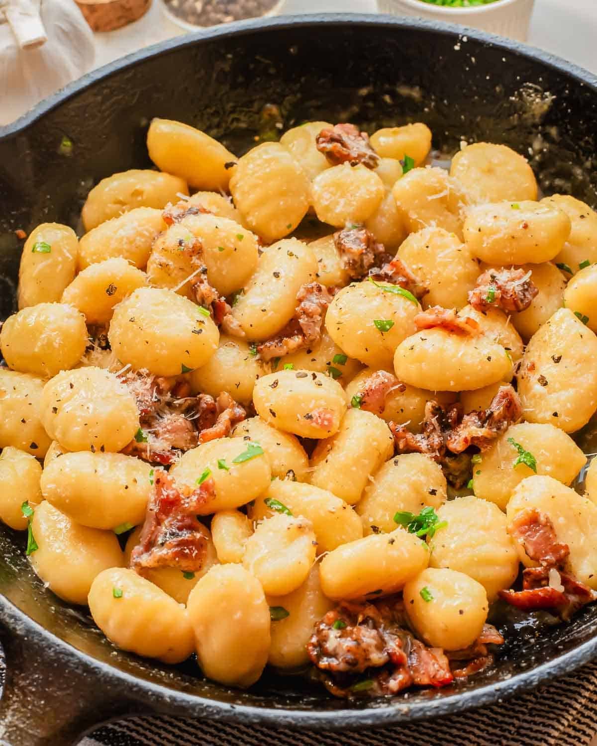 A skillet filled with creamy gnocchi mixed with pieces of crispy bacon, garnished with chopped parsley and grated cheese.