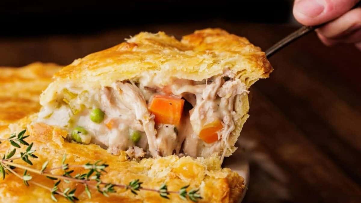 A close-up of a golden, flaky pot pie being served, showing a creamy filling of chicken, carrots, peas, and gravy; fresh thyme is visible in the foreground.