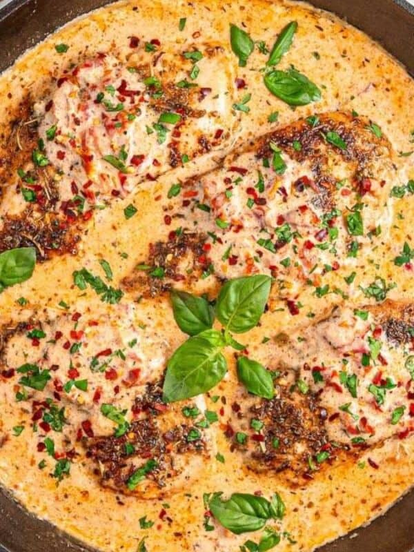 A cast-iron skillet with creamy Tuscan chicken breasts in a rich sauce, garnished with fresh basil, chopped parsley, and sun-dried tomatoes, sitting on a white marble surface.