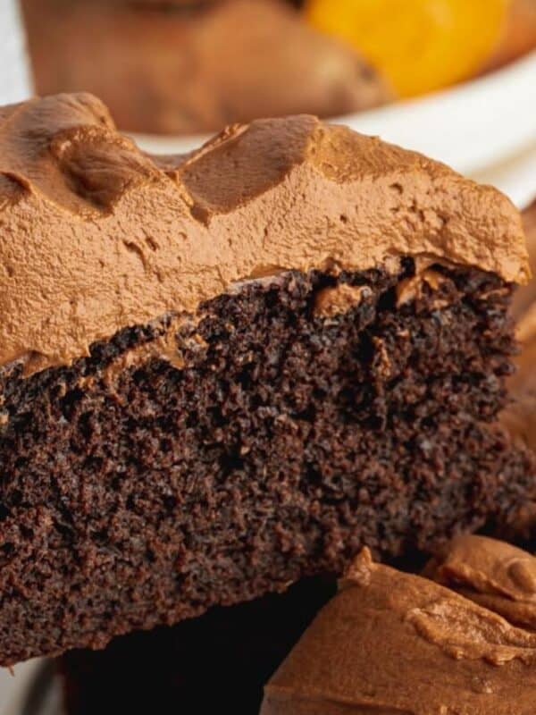 A close-up of a slice of moist chocolate cake with thick, creamy chocolate frosting being lifted from the whole cake, which is covered in swirls of the same frosting.