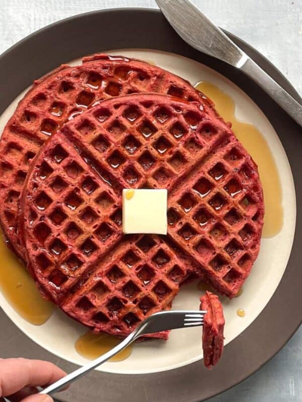 A hand holds a fork with a bite-sized piece of red waffles, topped with a square of butter and drizzled with syrup, on a white plate with a brown rim and cutlery on the side.